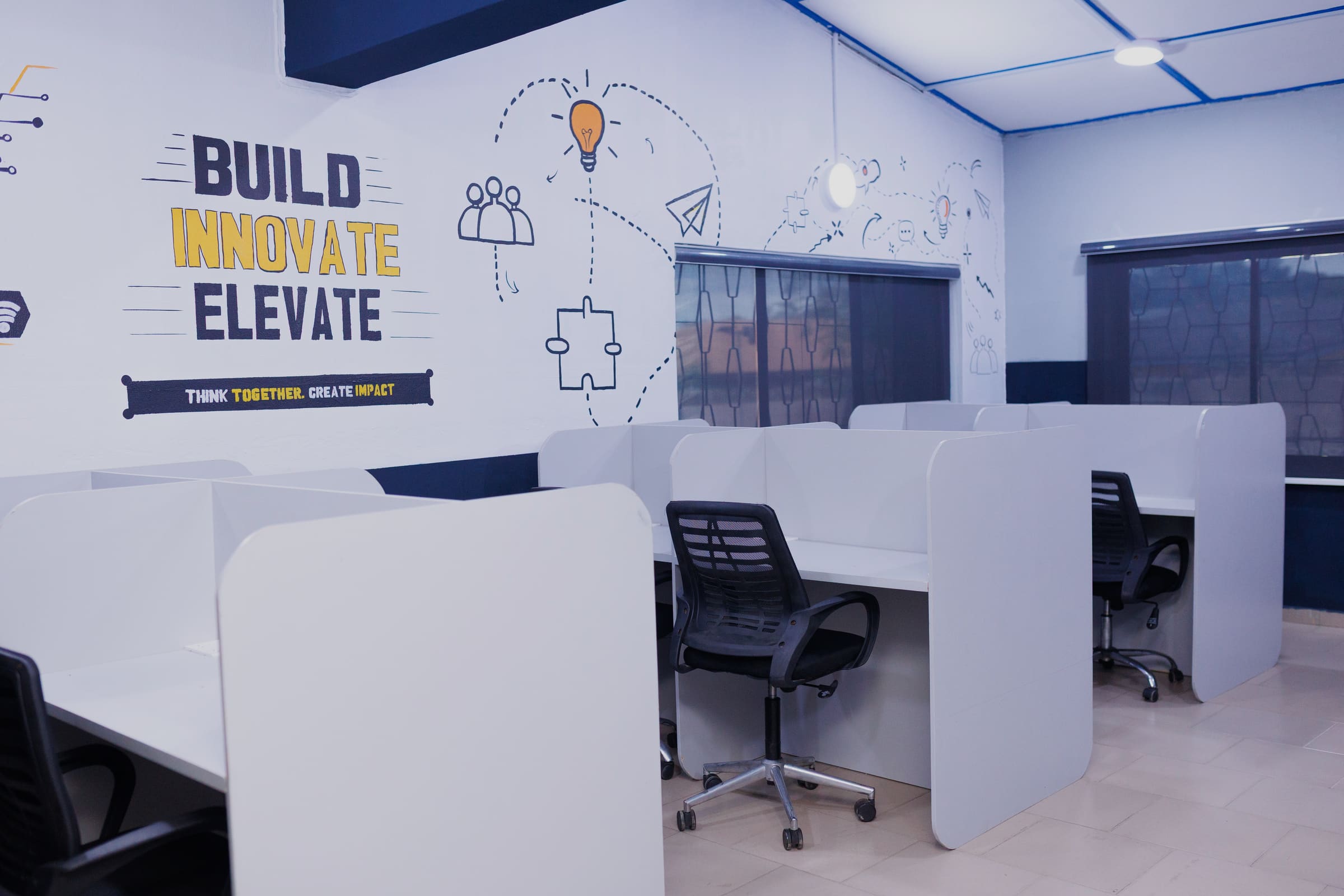 Desks divided by tall white partitions into individual enclosed bays, facing a mural reading "Build. Innovate. Elevate."