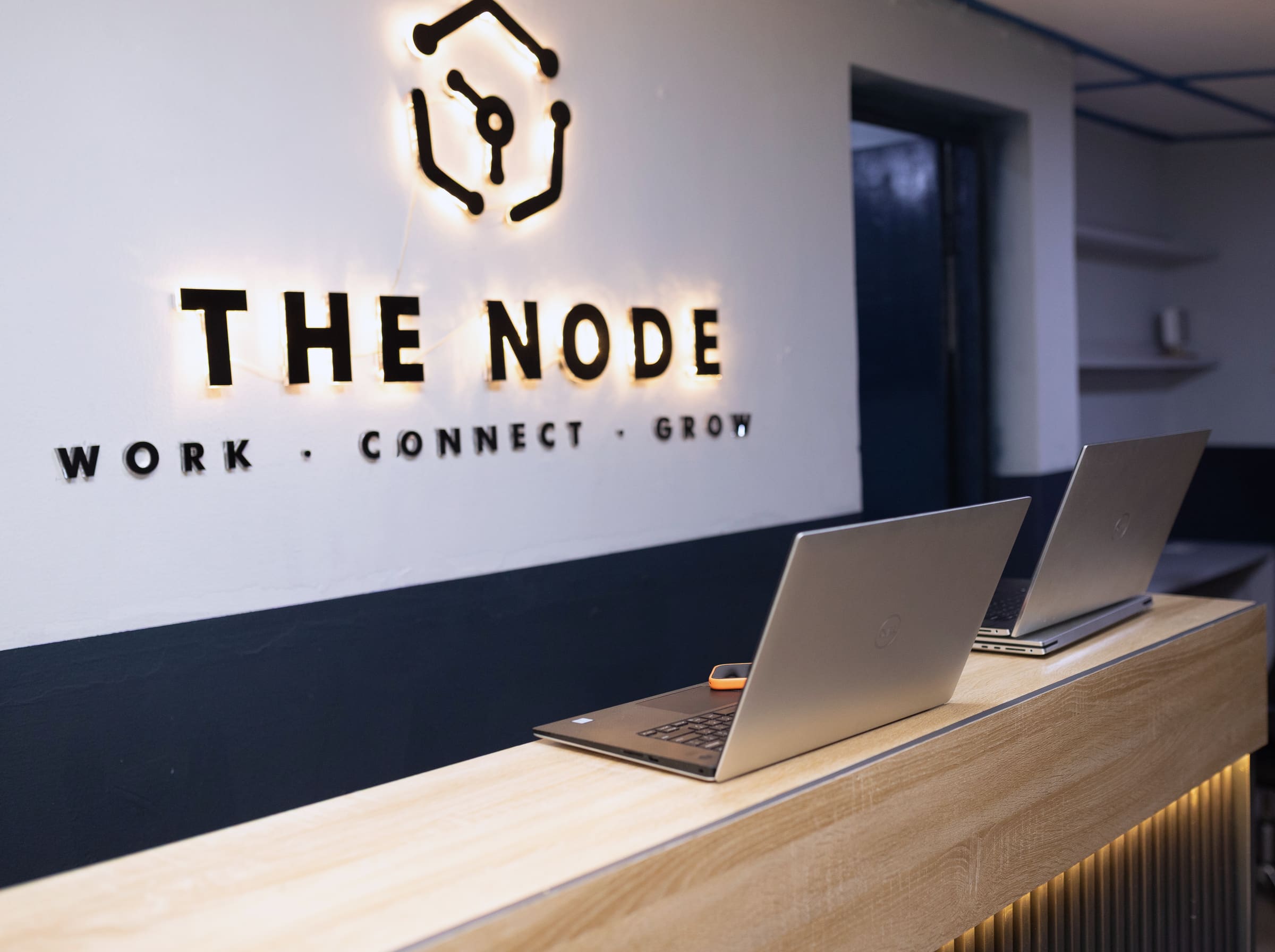 The reception desk at The Node Ibadan: a backlit logo above a wood counter, with two laptops on it.