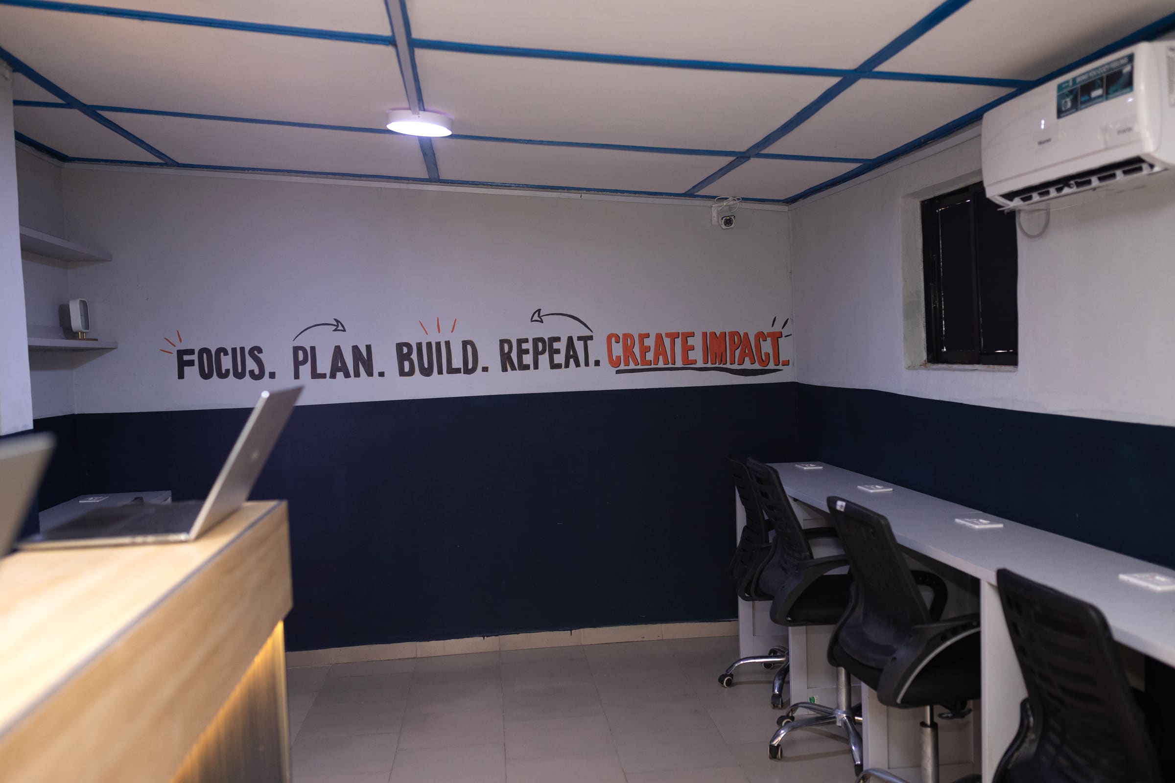 The open-plan room: a wall-length white desk with chairs along it, beneath a mural reading "Focus. Plan. Build. Repeat. Create Impact."